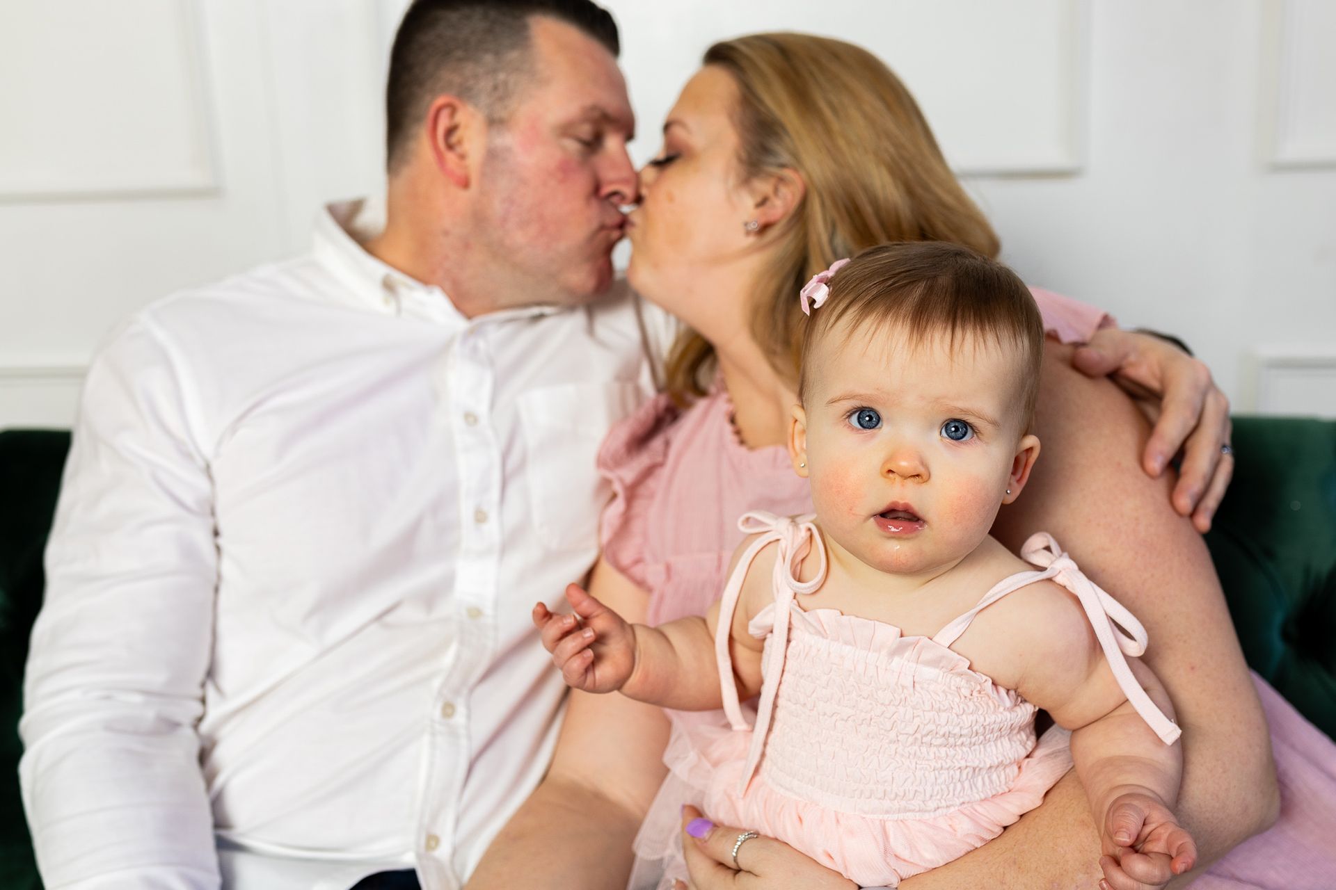 Family sitting on a couch, parents kissing while holding a baby in pink clothes - family photos