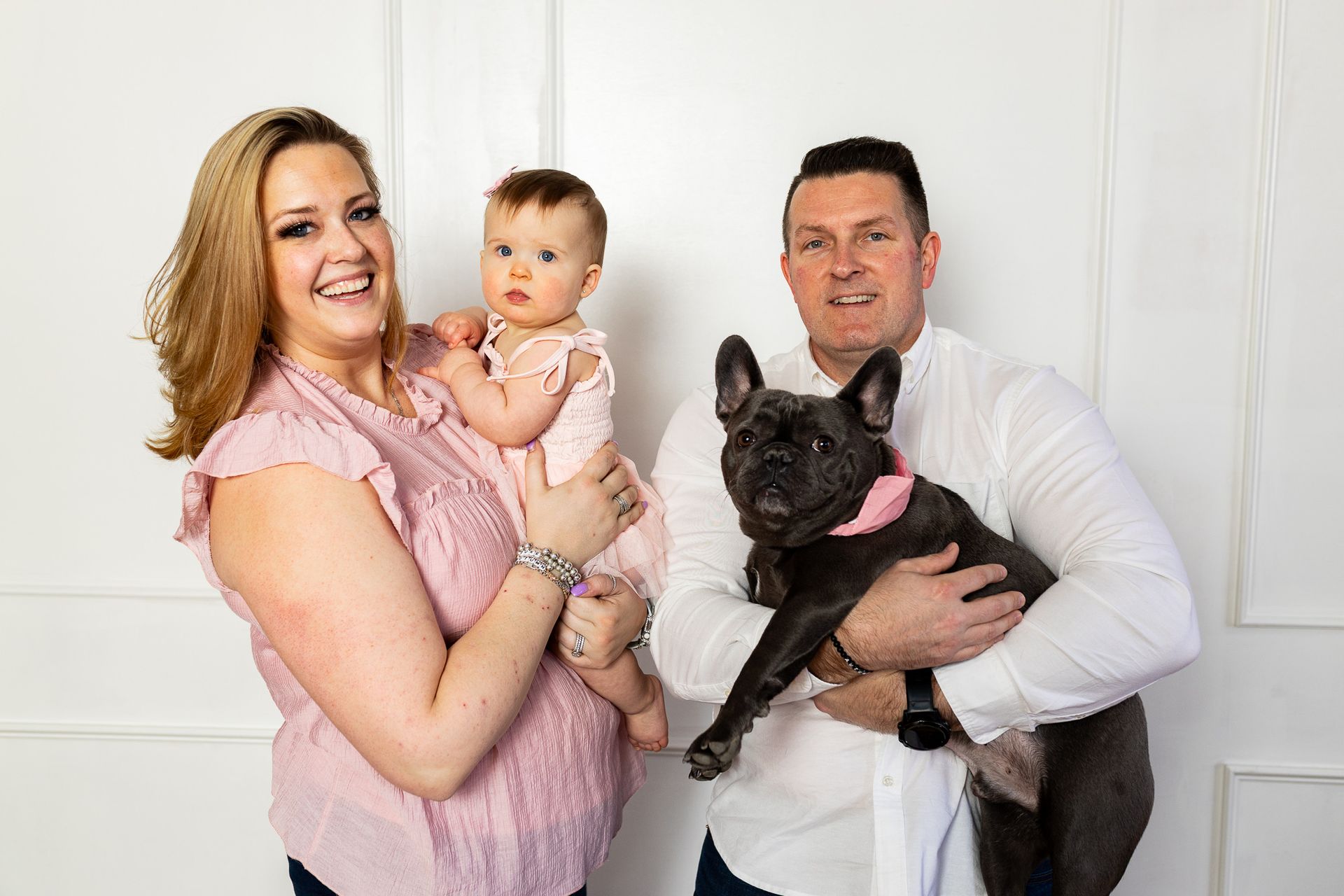 Smiling adults holding a baby and a black French bulldog against a white backdrop - family photos