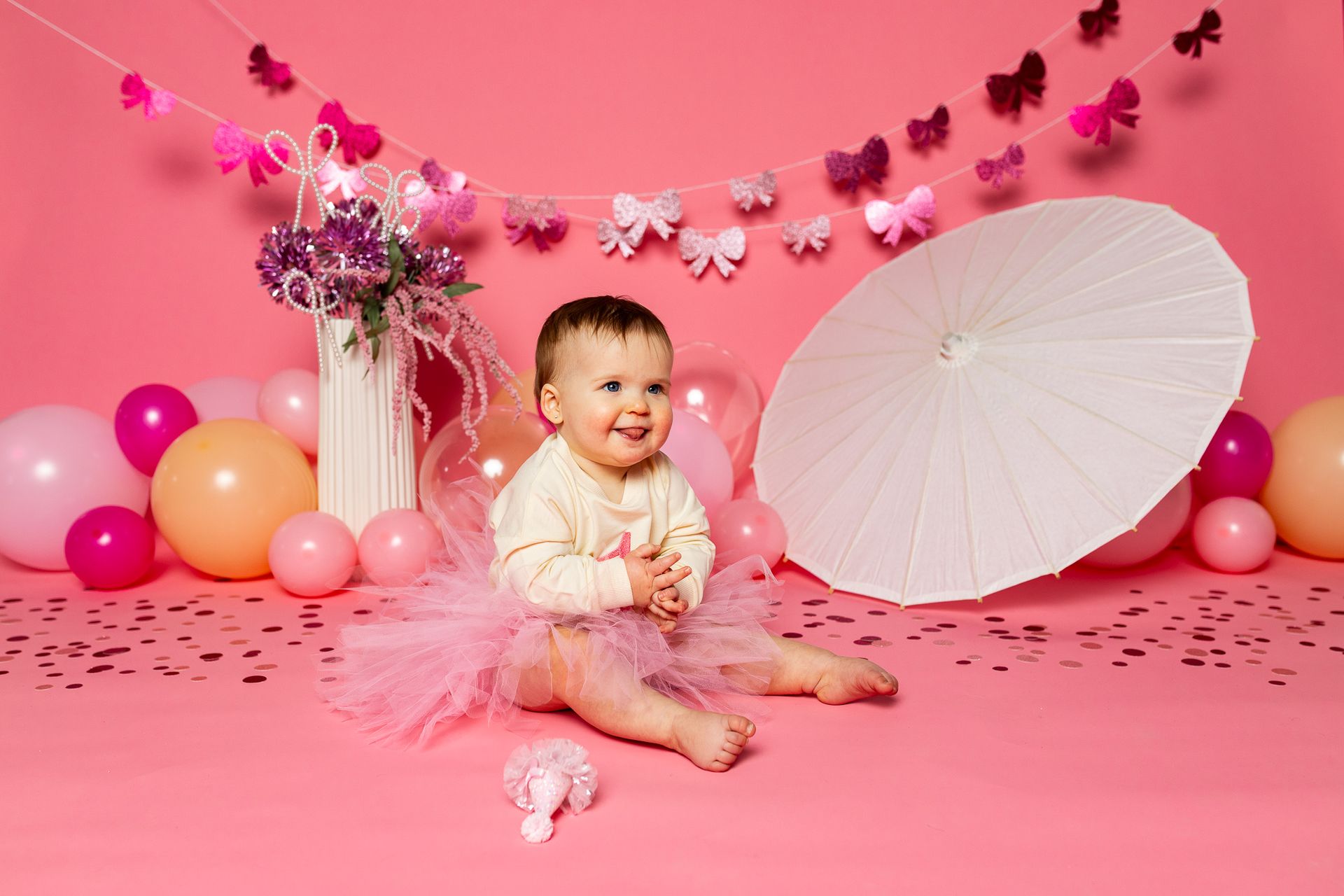 Baby sitting on pink floor amid balloons, flowers, and a white parasol in a pink cake smash setup