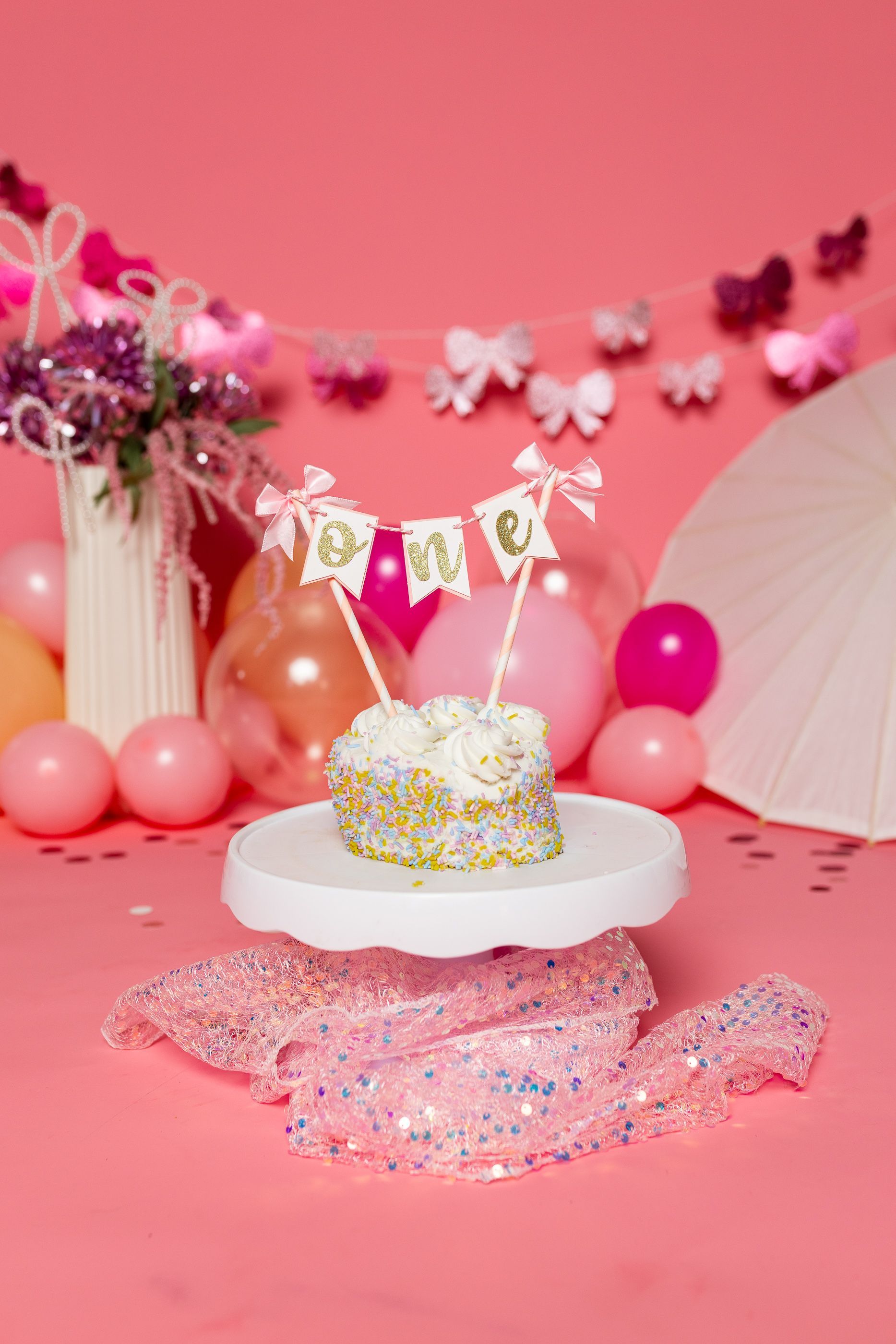 Baby sitting on pink floor amid balloons, flowers, and a white parasol in a pink cake smash setup
