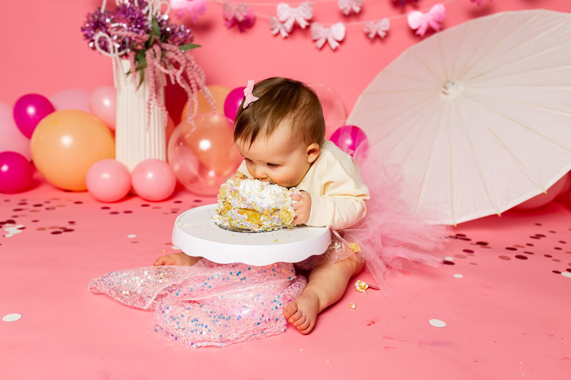 Baby sitting on pink floor amid balloons, flowers, and a white parasol in a pink cake smash setup