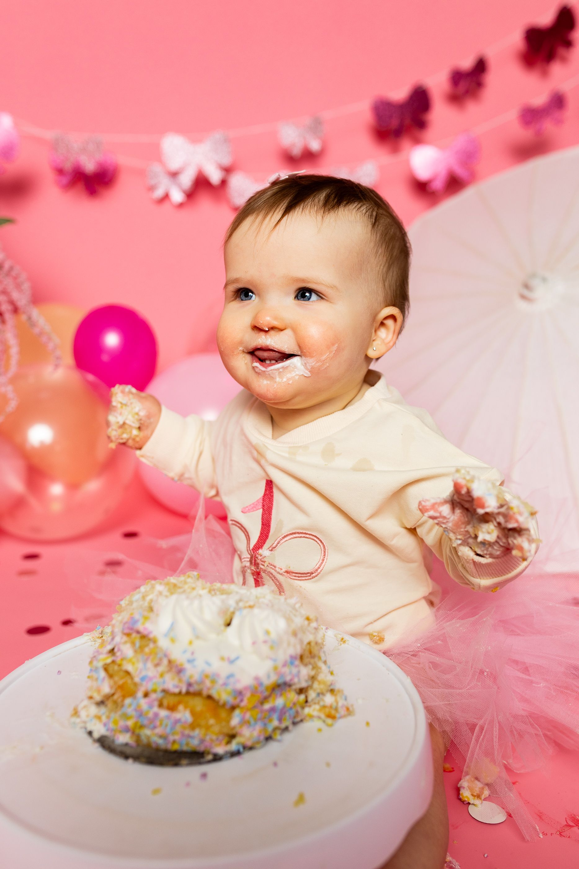 Baby sitting on pink floor amid balloons, flowers, and a white parasol in a pink cake smash setup