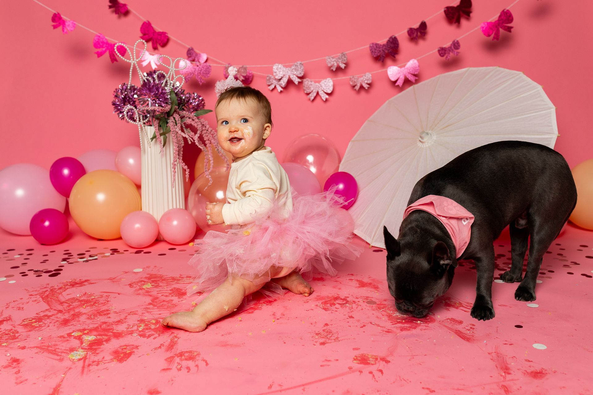 Baby sitting on pink floor amid balloons, flowers, and a white parasol in a pink cake smash setup