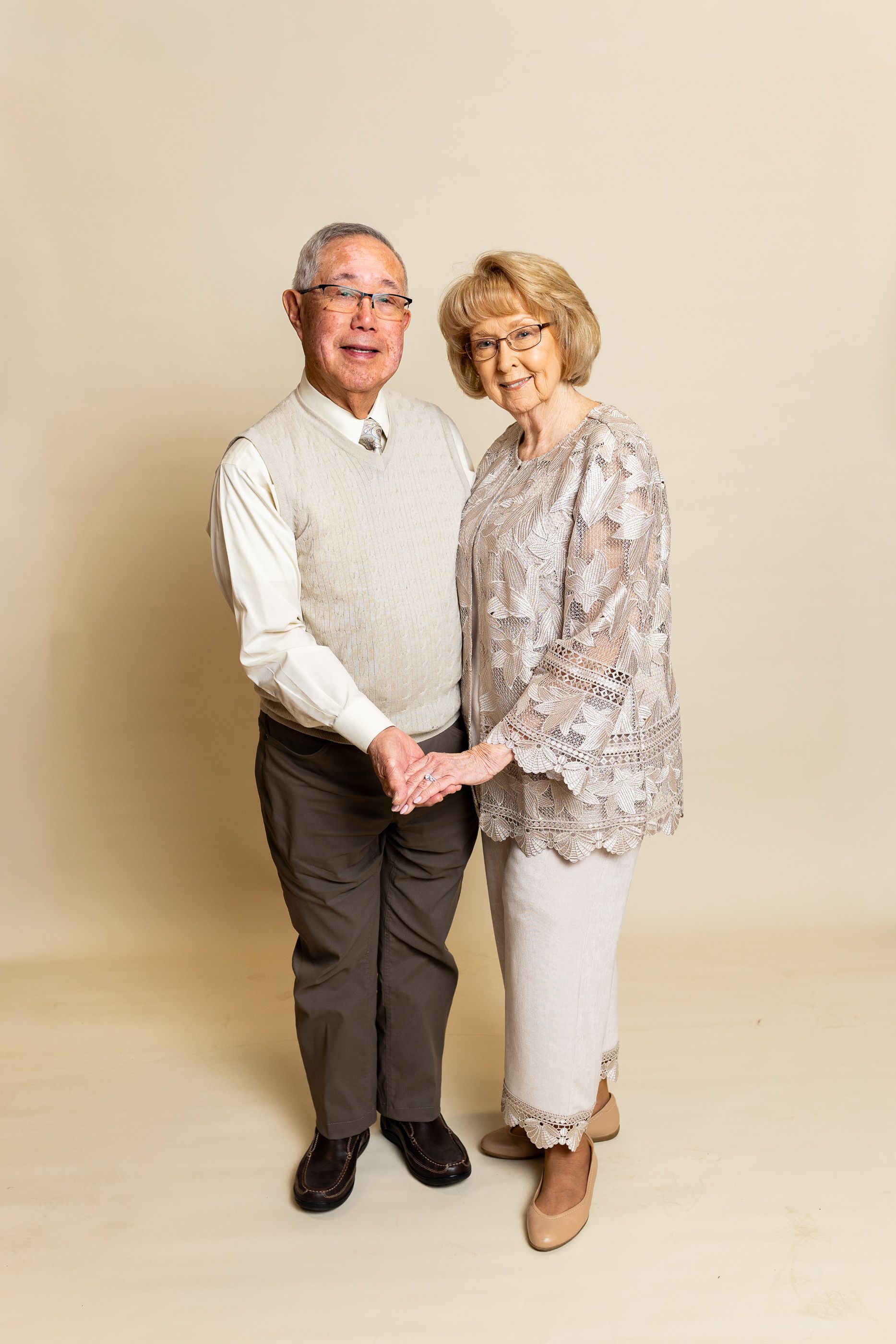 Two people standing together against a beige backdrop, holding hands and smiling at the camera. - family photos
