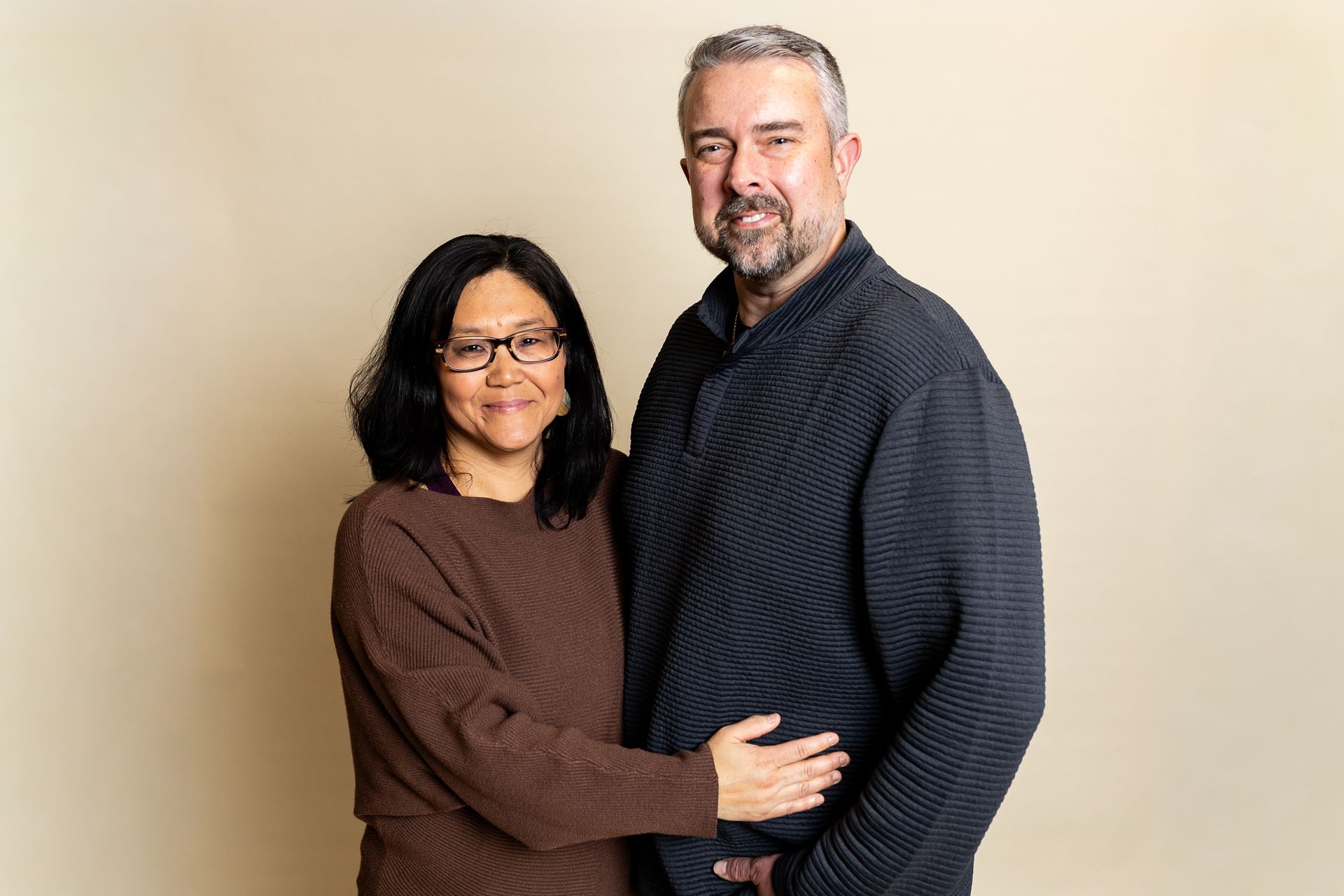 Two people standing together against a beige wall, smiling and embracing - family photos