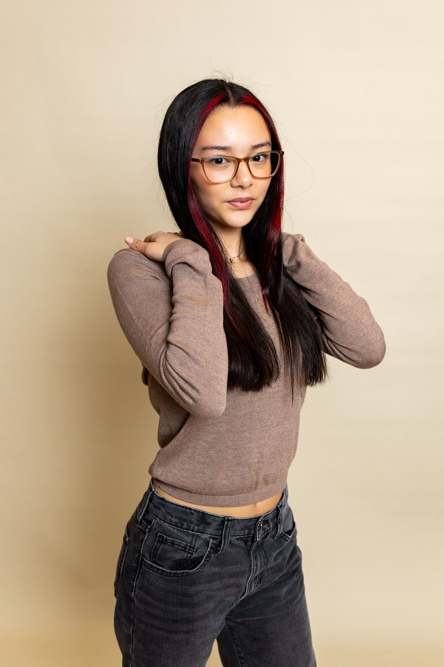Woman standing against a beige background, wearing glasses, a brown sweater, and jeans with hands near her shoulders. - family photos