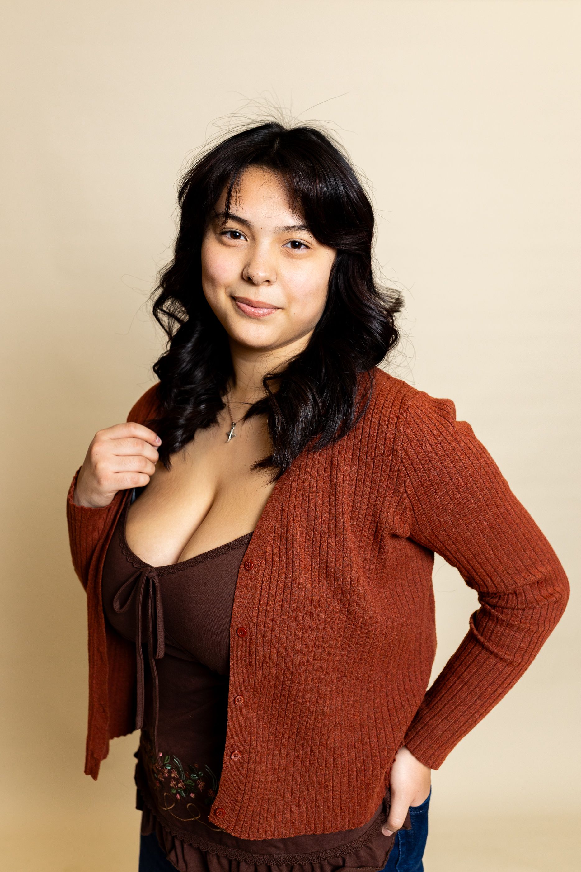Woman standing against a beige background, wearing a rust cardigan and brown top, with one hand on her hip. - family photos