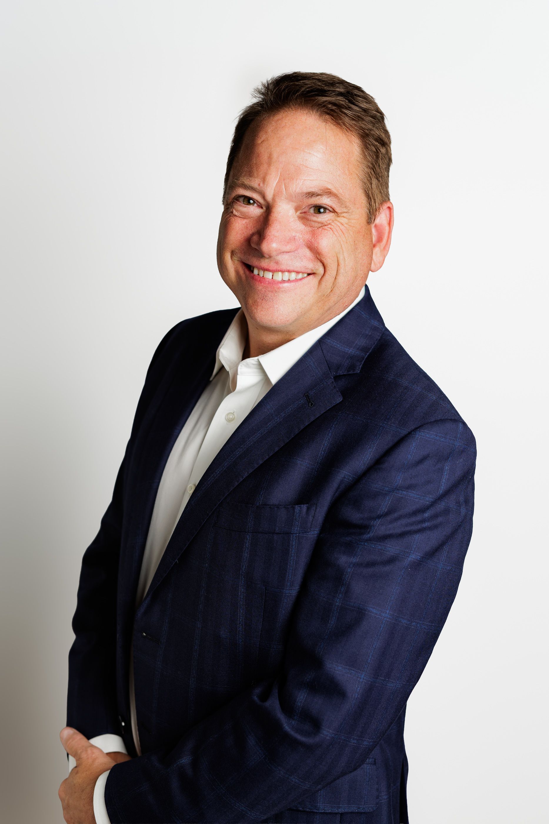 Smiling man in a navy blazer and white shirt, standing against a light background. - headshot session