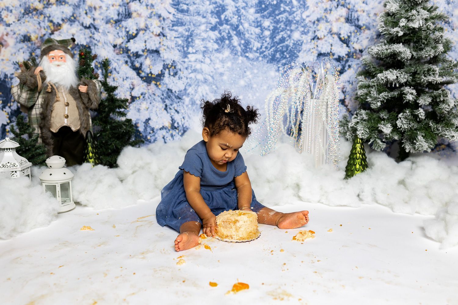 baby standing in a winter themed studio set with snowy background, mini christmas trees, and santa for cake smash session