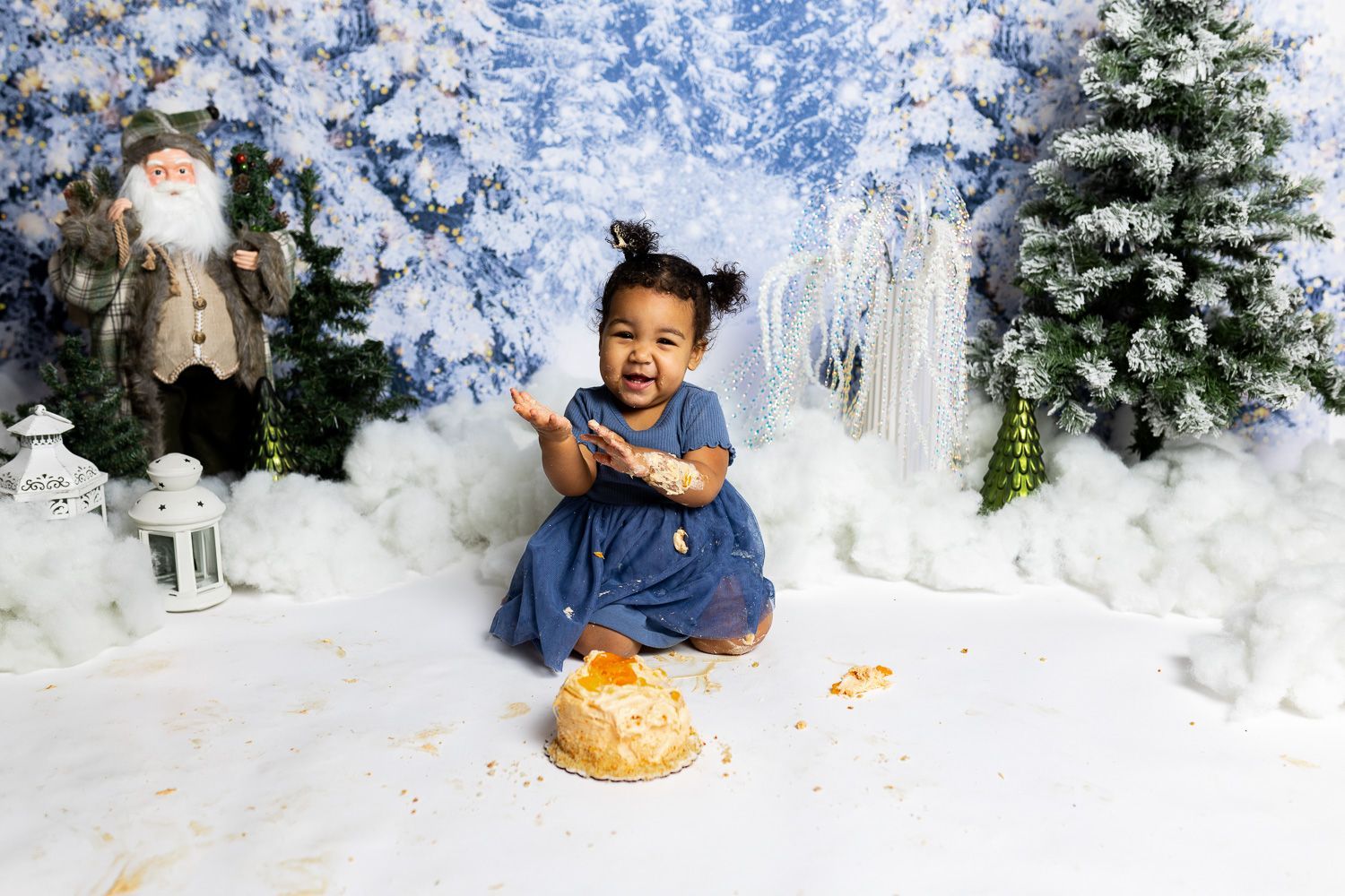 baby standing in a winter themed studio set with snowy background, mini christmas trees, and santa for cake smash session