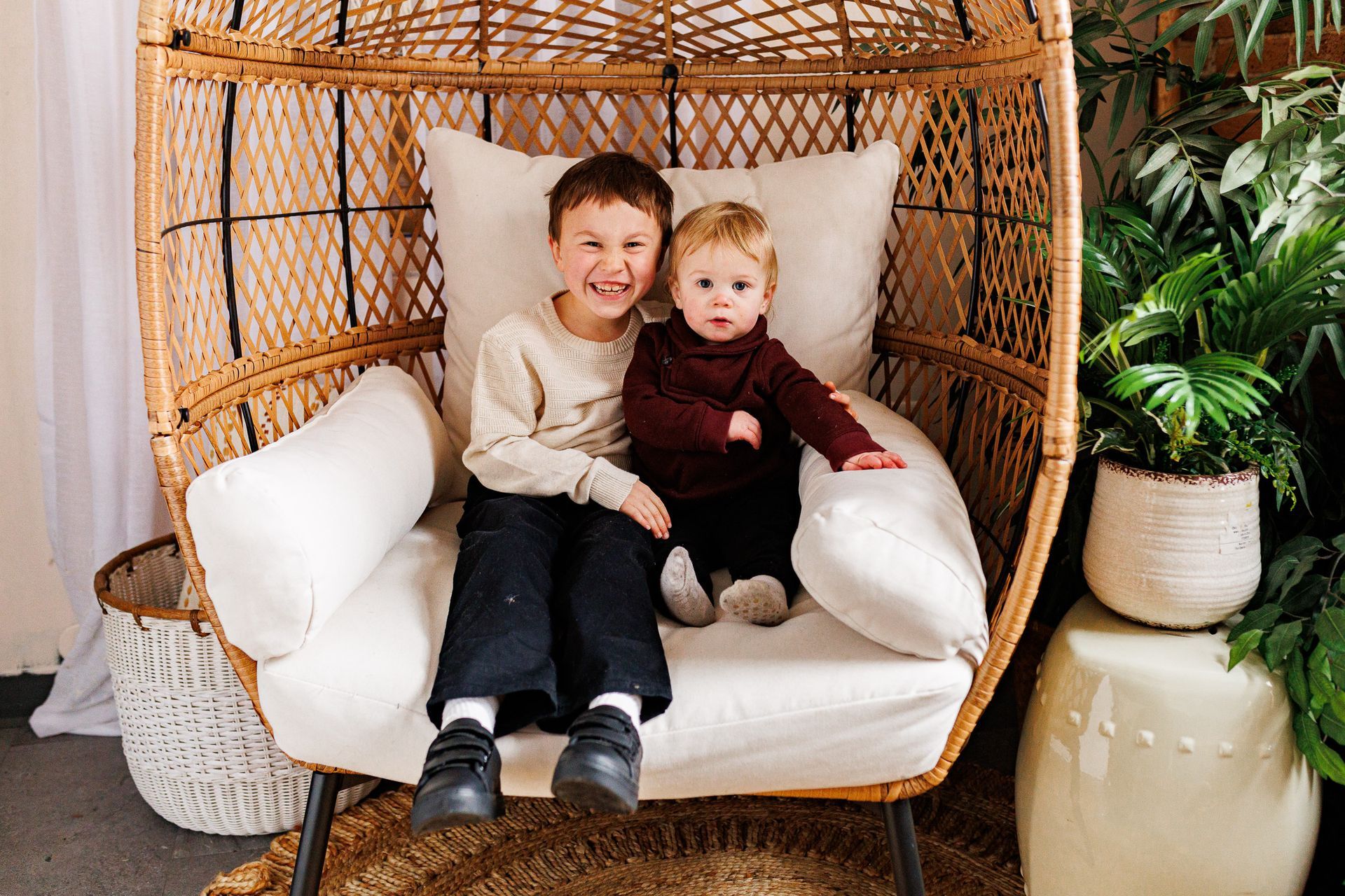 Two children smiling on a wicker chair with white cushions, surrounded by indoor plants. - family photos
