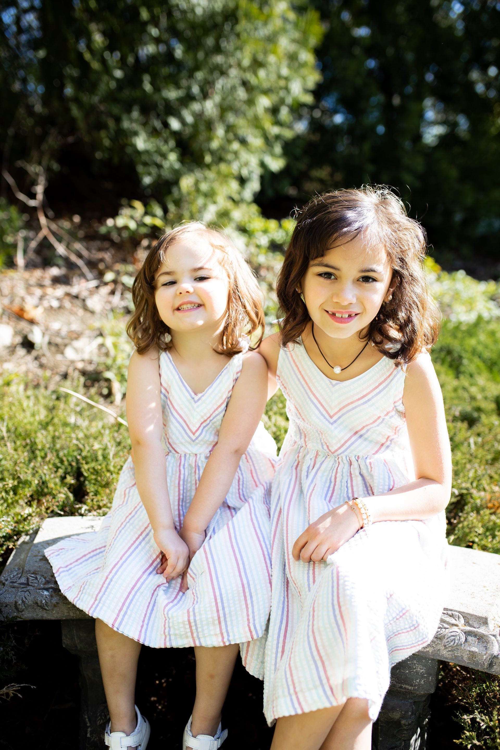 Two children in white striped dresses sitting on a stone bench outdoors in sunlight - family photos
