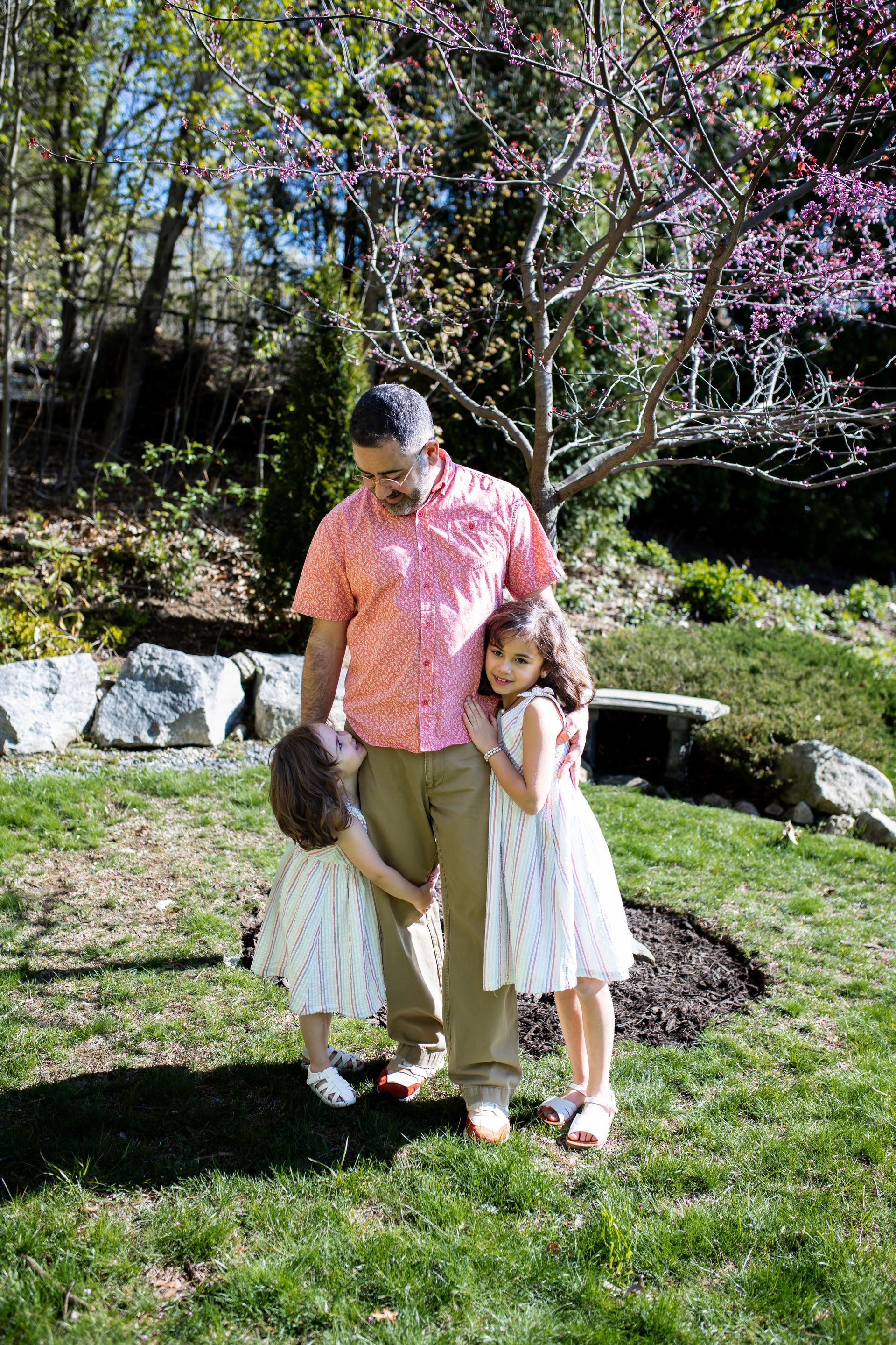 Three people standing on grass in a garden, with flowering trees and rocks behind them. - family photos