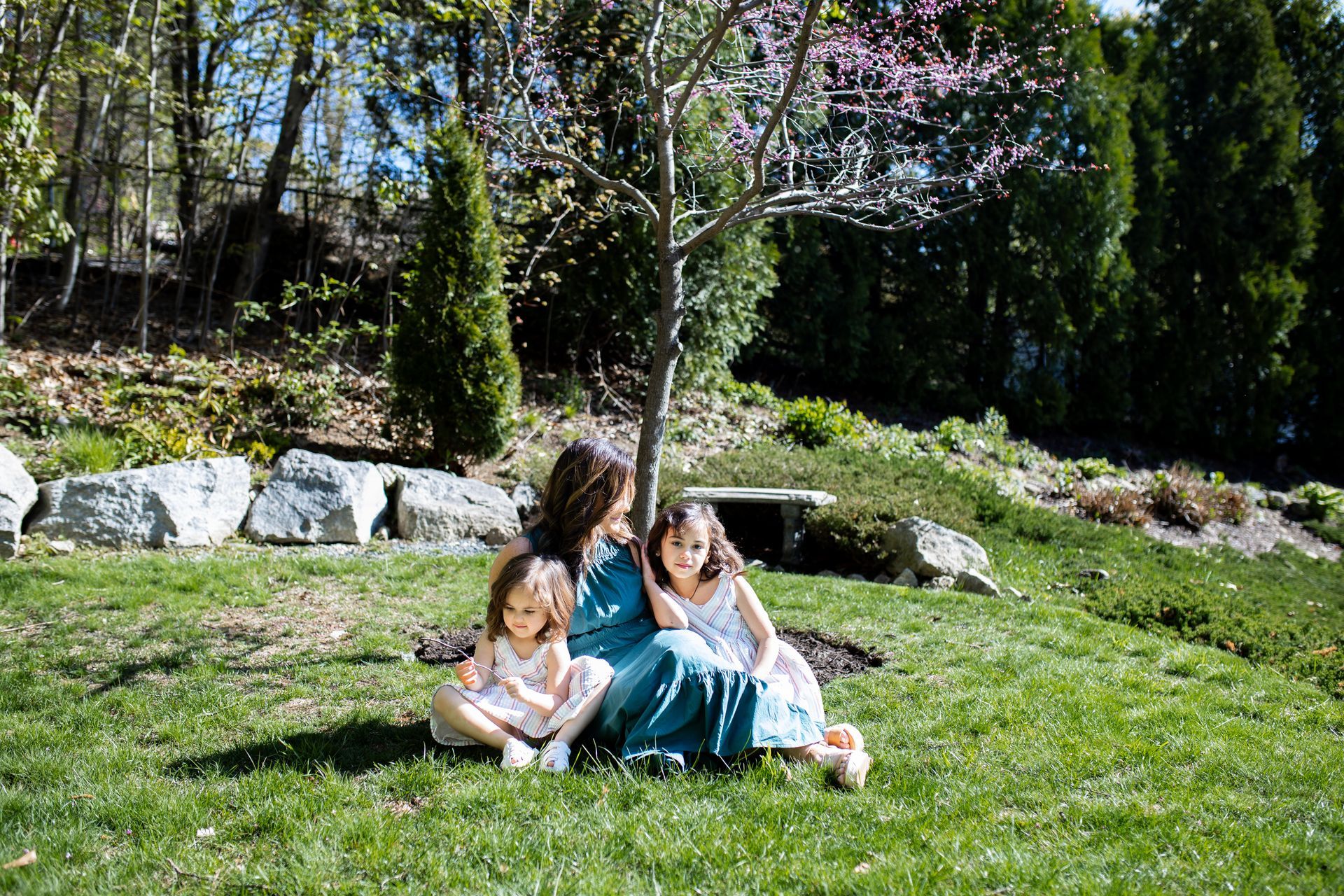 Two children sitting on grass under a tree in a sunny park, one in blue dress, one in light clothes - family photos
