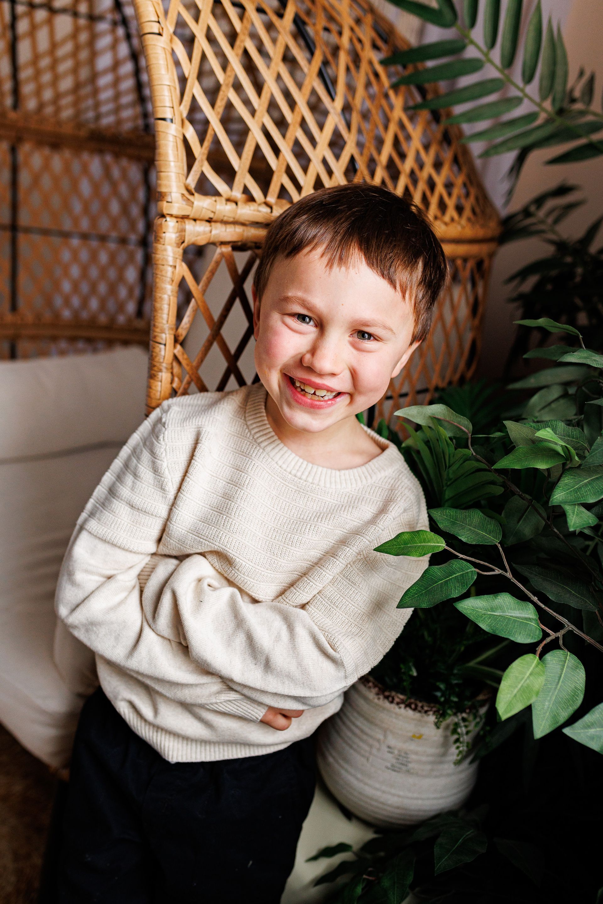 Smiling child in a cream sweater standing indoors beside a leafy plant and woven room divider - family photos
