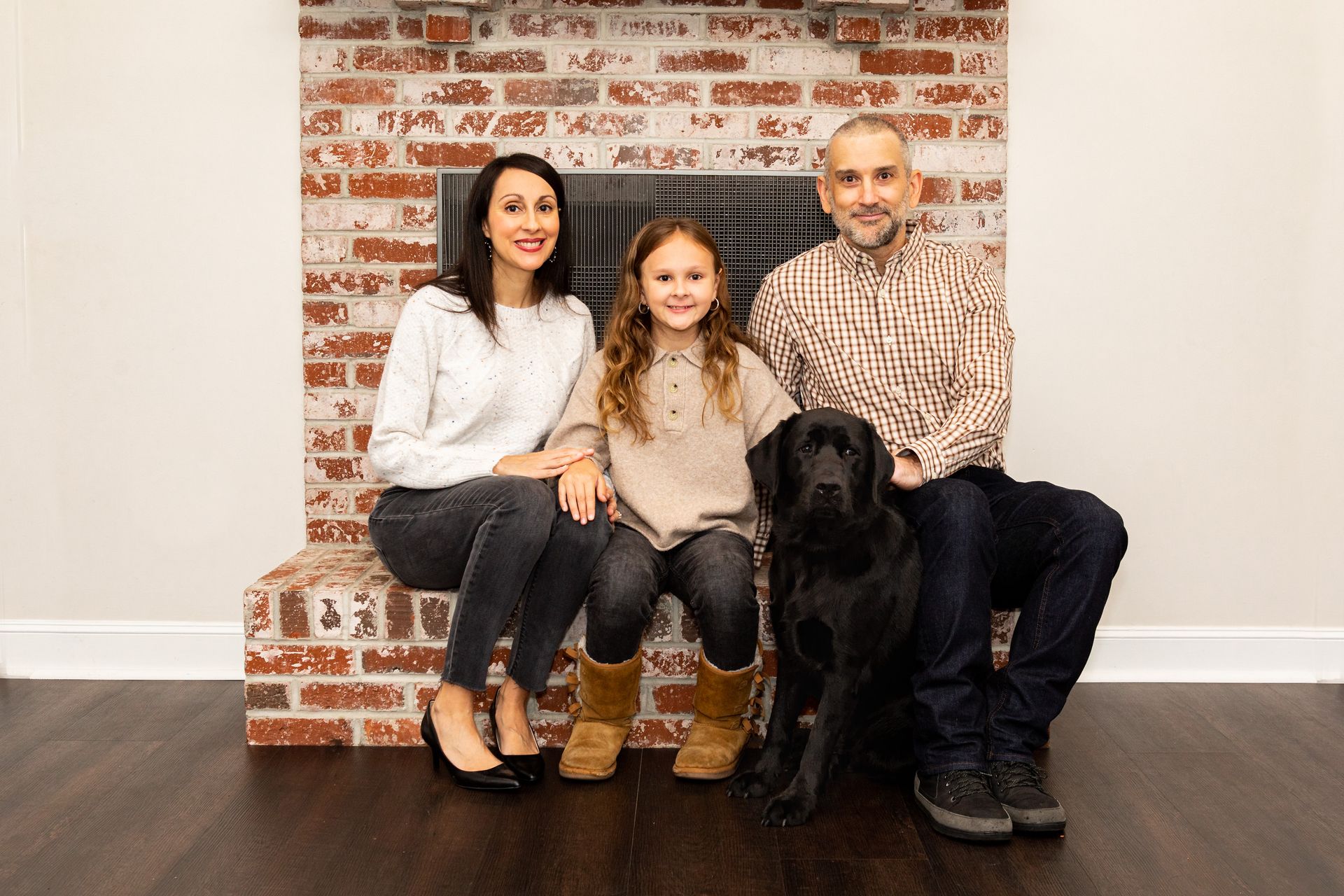 Family seated on a bench with a black dog in front, against a brick wall backdrop - family photos