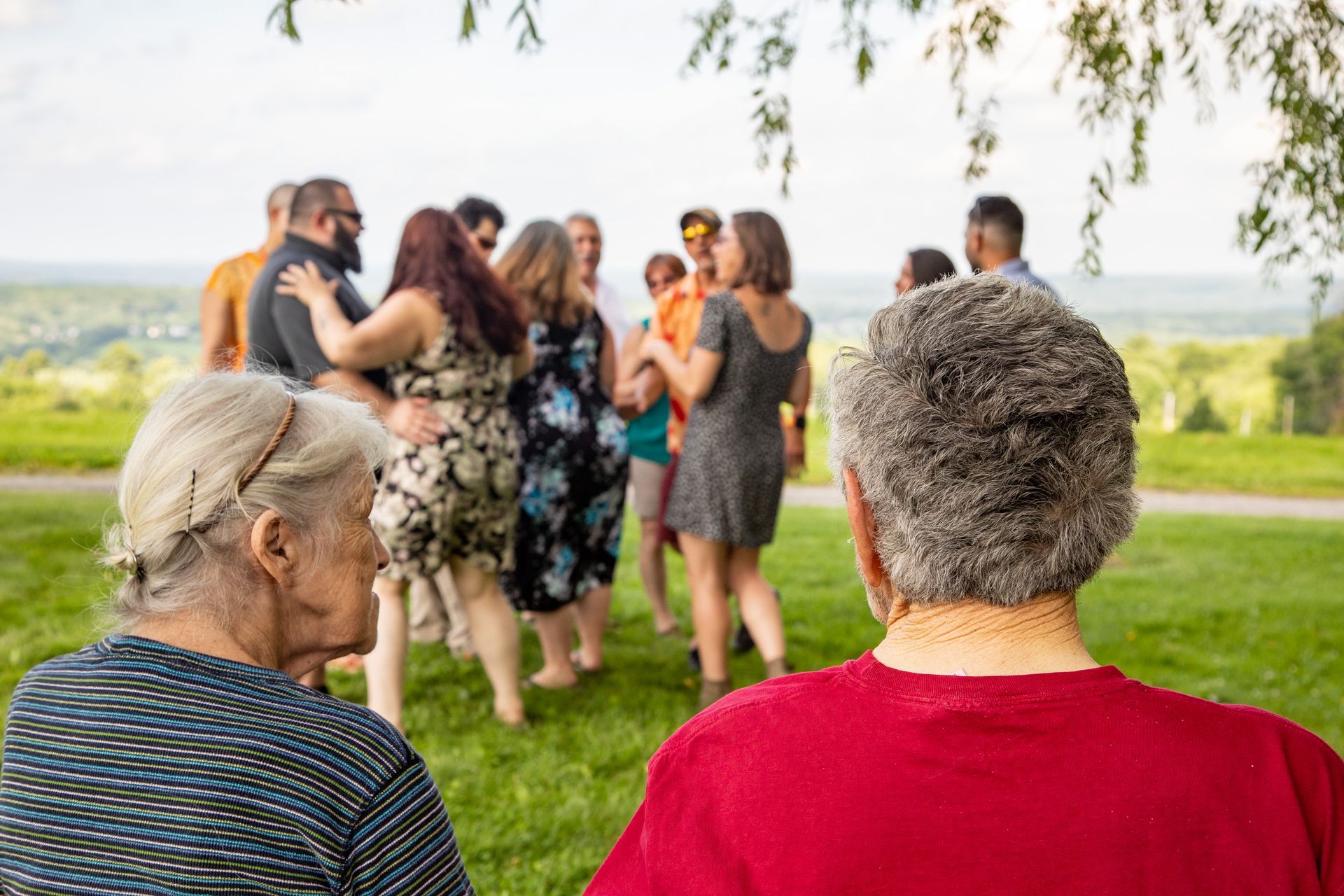 Two people watching a small group chatting outdoors on a grassy hill overlooking a landscape - family photos
