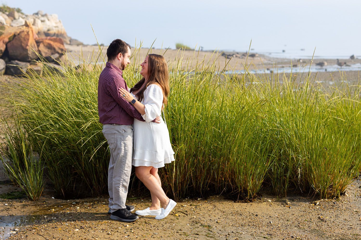 Couple embracing while on the beach in front of long grass during engagement session
