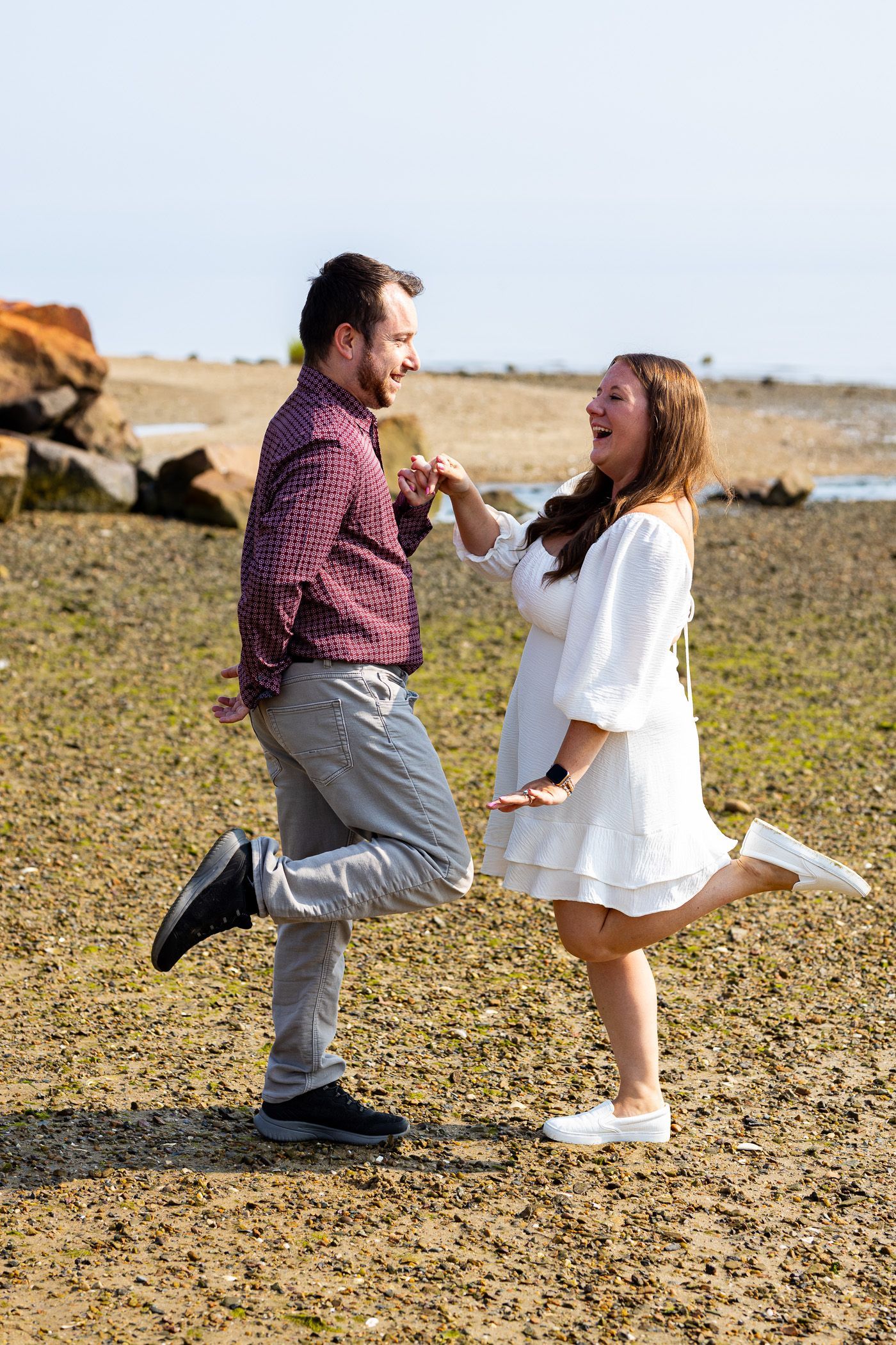 Couple laughing at each other while kicking up 1 foot on the beach during engagement session