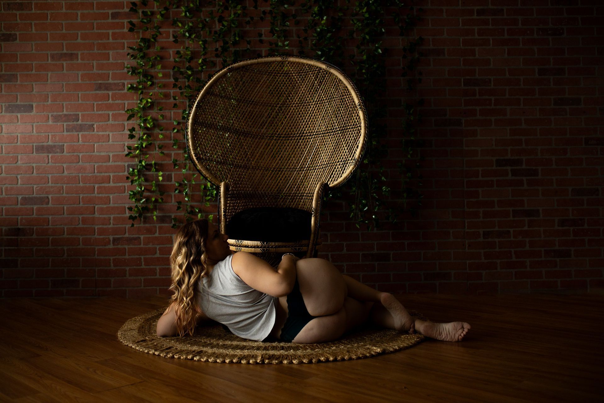Woman posing in lingerie for boudoir session lying on floor in front of a peacock chair and vines