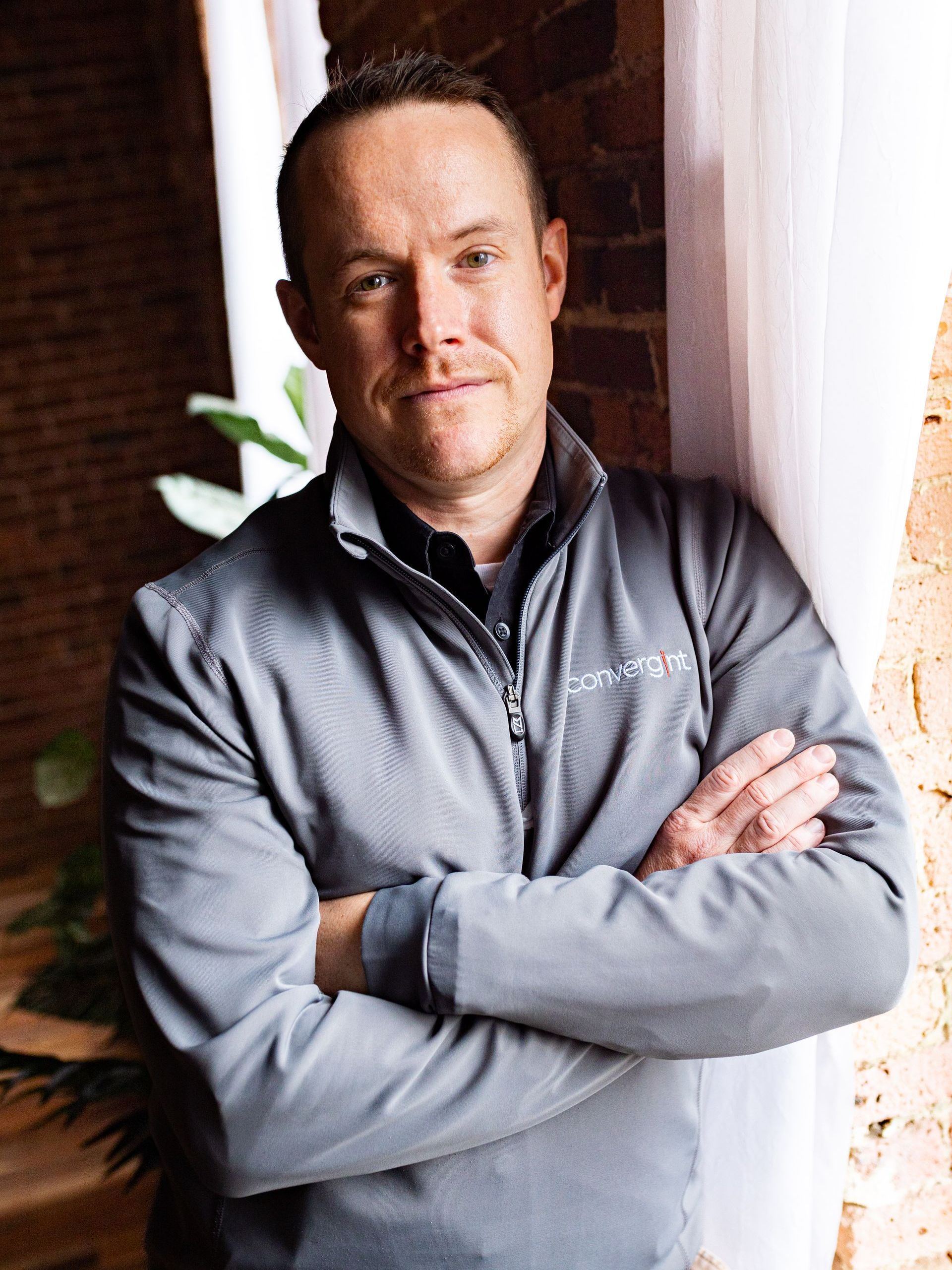 Photography MA -A man in a grey jacket is standing with his arms crossed in front of a window.