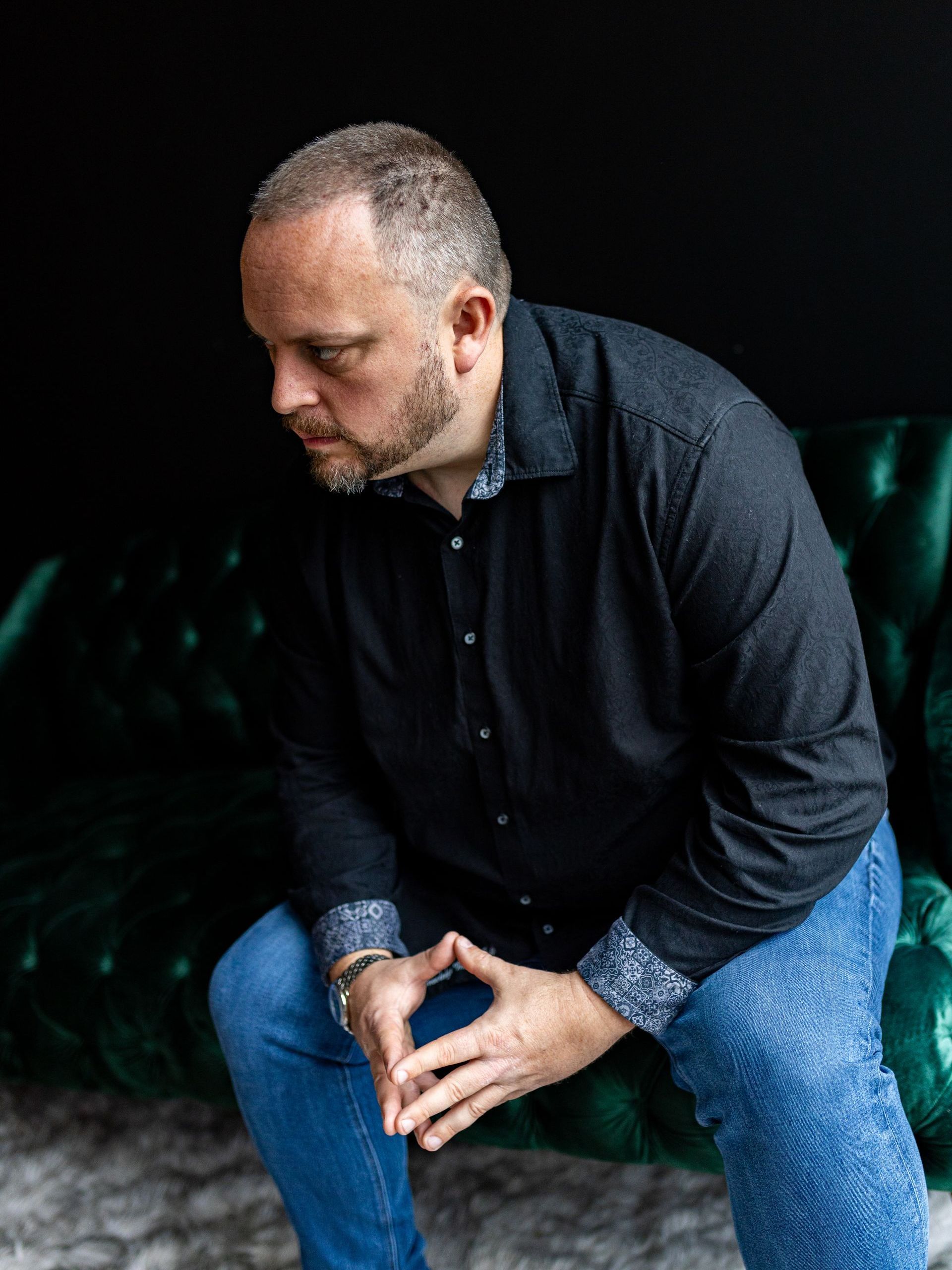 Photography MA -A man in a black shirt and blue jeans is sitting on a green couch.