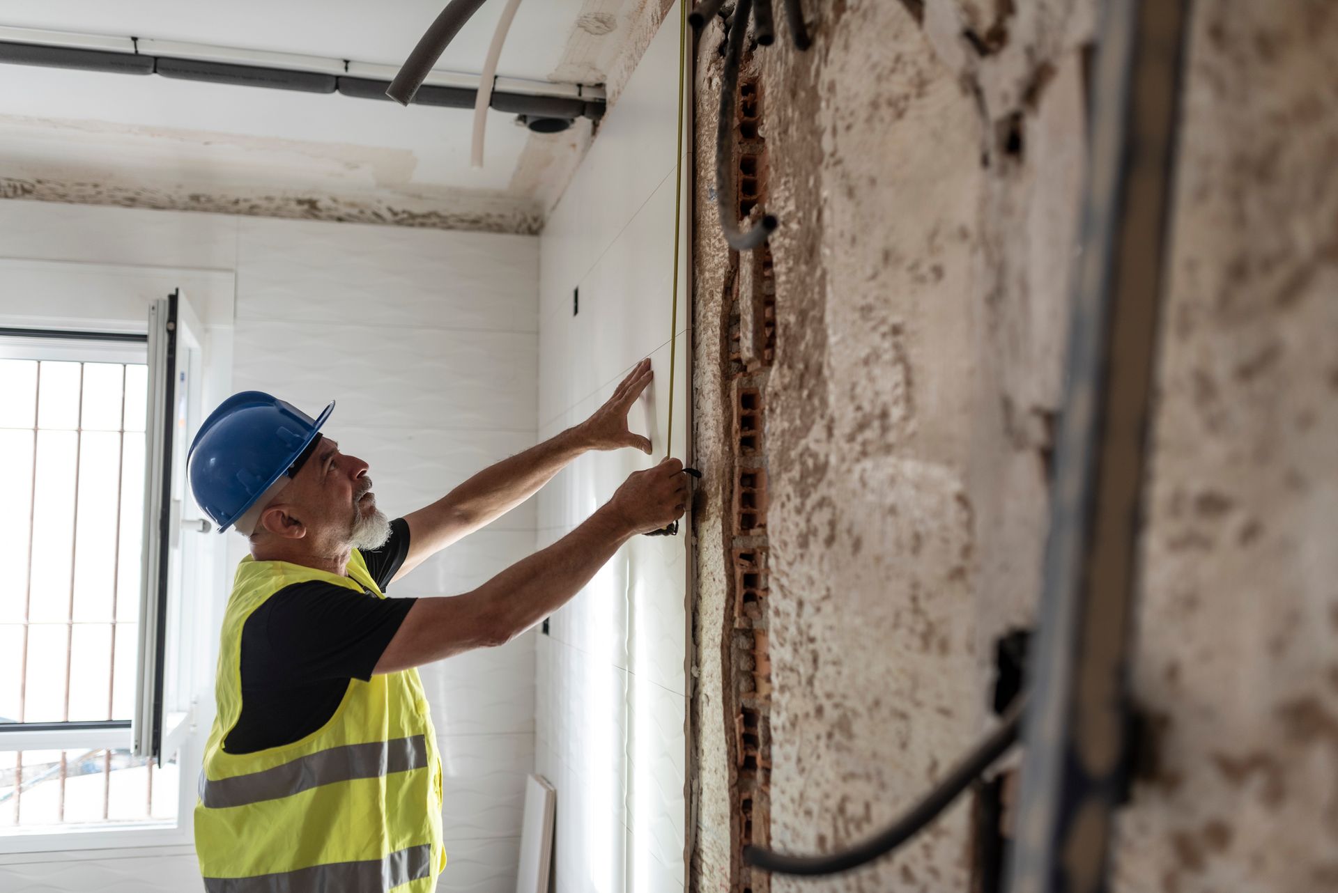 A man wearing a hard hat and safety vest is working on a wall.