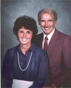 A smiling man and woman pose together, woman wearing a blue shirt and pearl necklace, man in a red suit and tie.