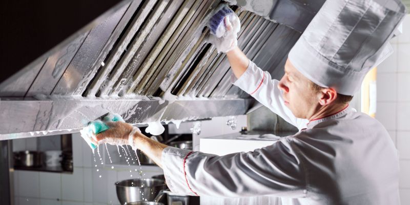 Chef cleaning a stainless steel kitchen hood with a spray bottle and cloth