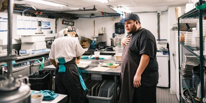 Two people in a commercial kitchen, one preparing food at a counter and one standing nearby.