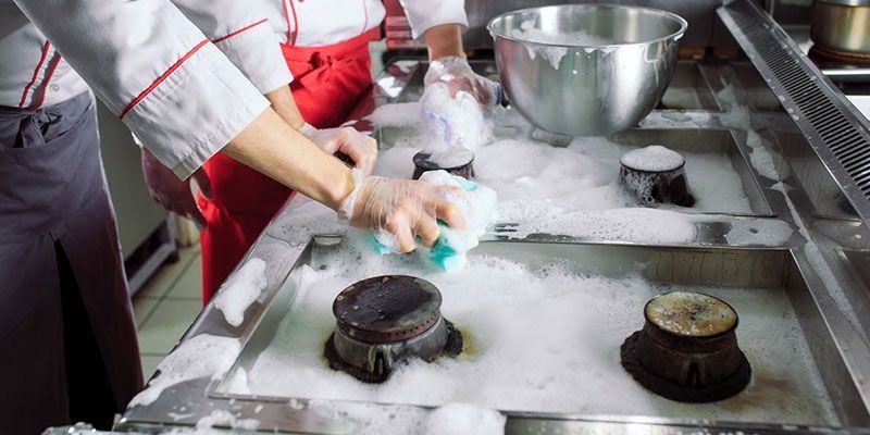 Worker cleaning a dusty stovetop in a kitchen with metal bowls and red apron visible
