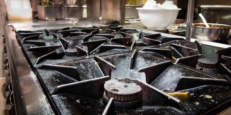 Row of black gas stove burners in a commercial kitchen, with metal pots and pans in the background