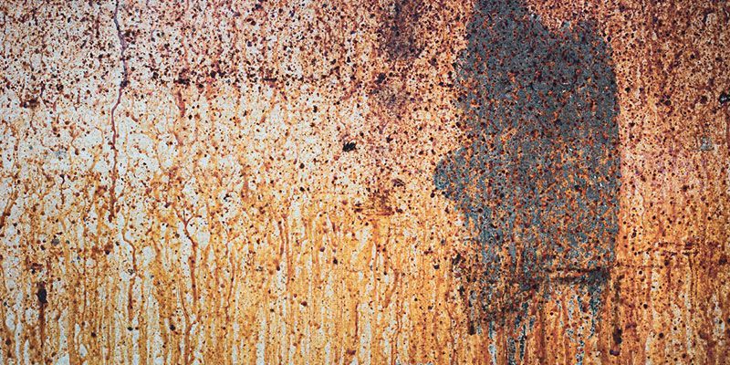 Weathered rusty metal wall with faded orange-brown rust streaks and a dark patch on the right