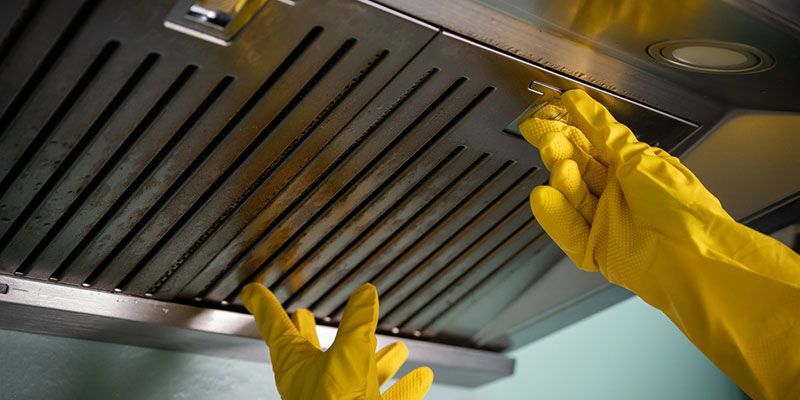 Gloved hands reaching up to clean a stainless steel kitchen range hood filter
