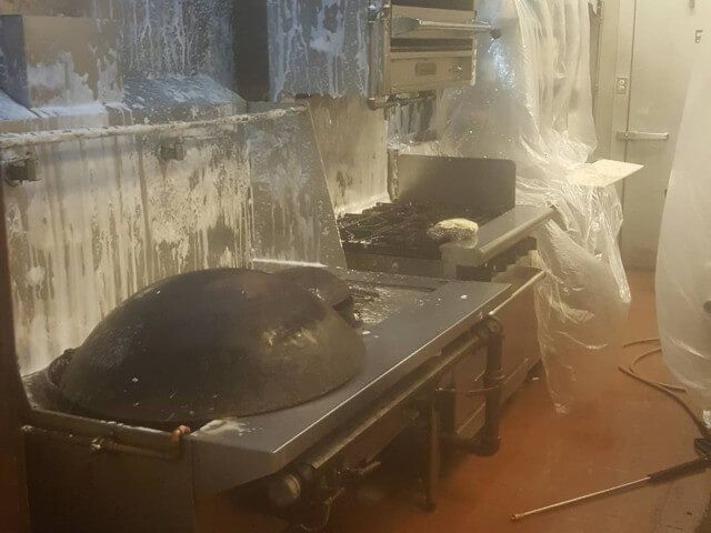 Industrial kitchen with a large black pan on a stove, covered in steam and grease splatter