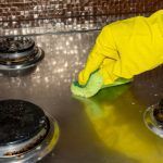 Yellow-gloved hand wiping a dirty stovetop with a green sponge