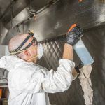 Worker in protective gear spray-coating a metal surface inside a reflective industrial chamber.