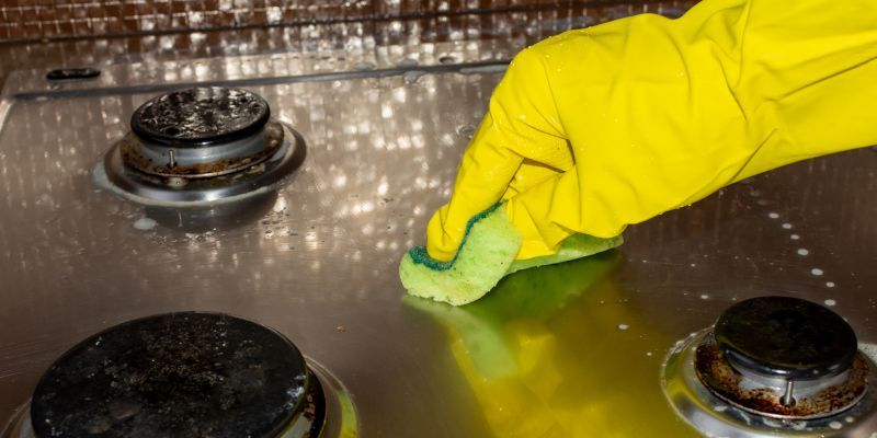 Gloved hand cleaning a stainless steel stovetop with a green cloth