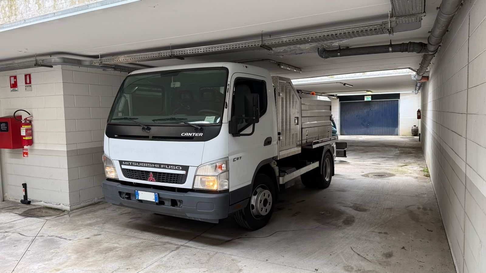 Un camioncino bianco con pianale ribassato è parcheggiato in un corridoio coperto di un garage con pareti in cemento e una saracinesca blu nelle vicinanze.