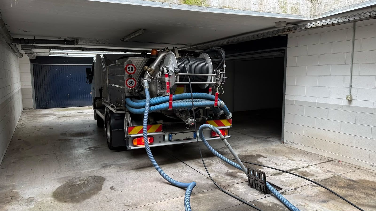 Camion dotato di tubo flessibile sotto un soffitto basso in cemento in un garage o tunnel stretto, con pavimento bagnato e cavi.