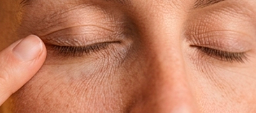 Close-up dos olhos fechados de uma pessoa com os dedos pressionando suavemente a área da pálpebra que tem linhas finas.