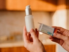 Mãos segurando um frasco de cosmético branco e uma pequena caixa retangular do produto em um ambiente interno aquecido.