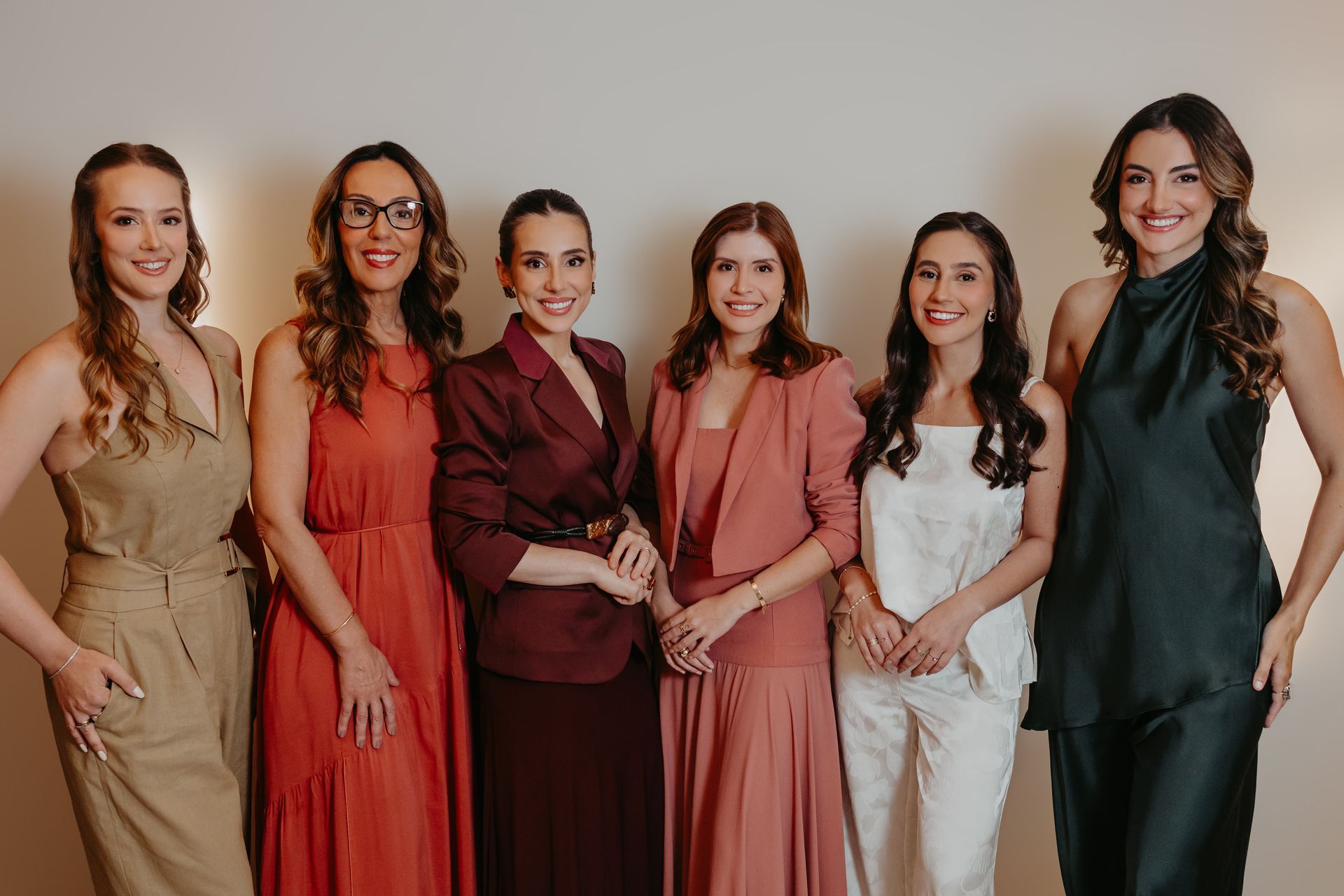 Seis mulheres, vestidas formalmente, posam lado a lado contra um fundo de estúdio em tons quentes.