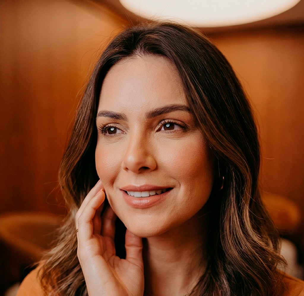 Mulher sorridente com longos cabelos castanhos em um interior aconchegante e com iluminação suave.