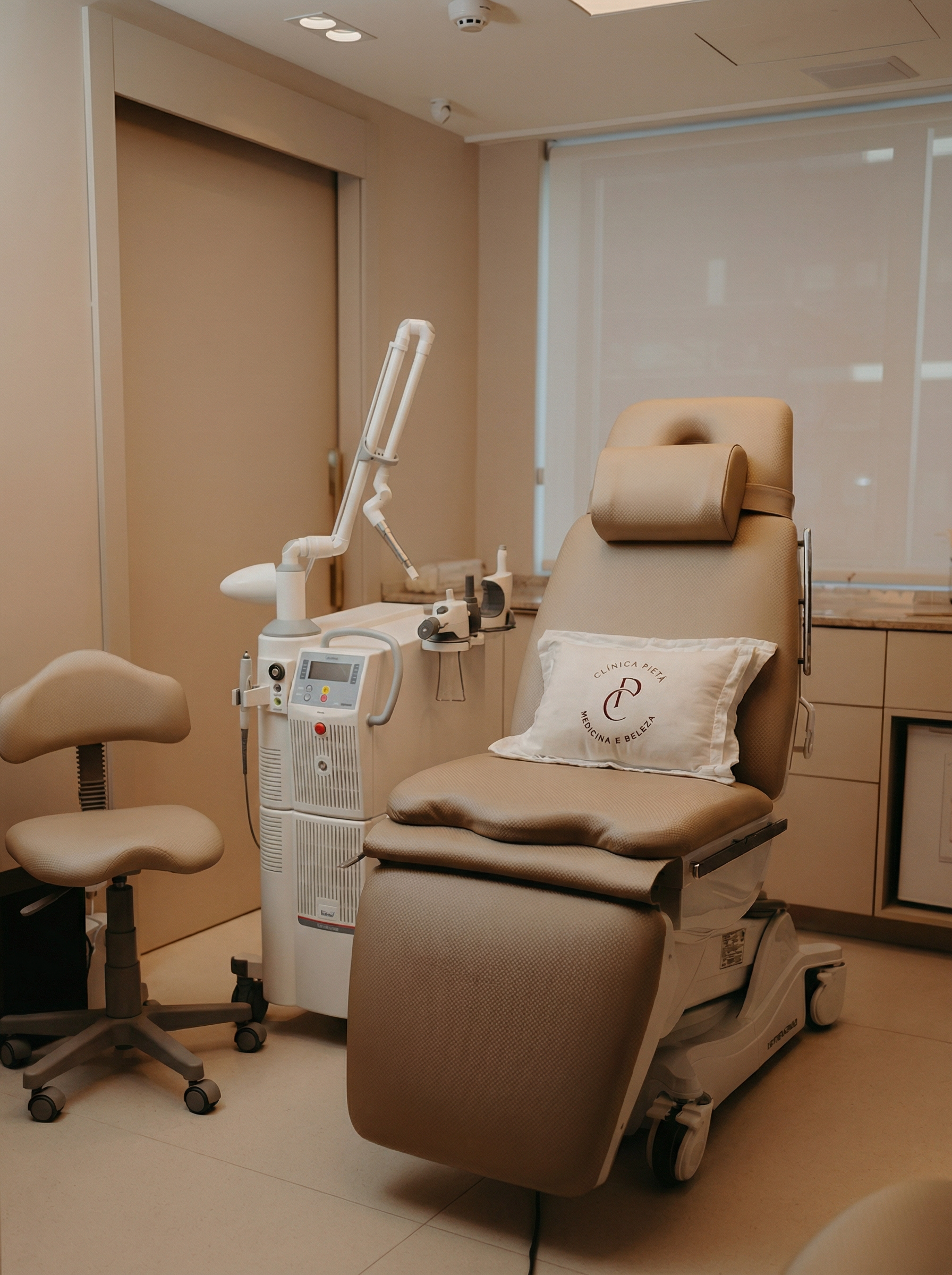 Sala de exame médico vazia, de cor bege, com cadeira de tratamento ajustável e equipamentos.