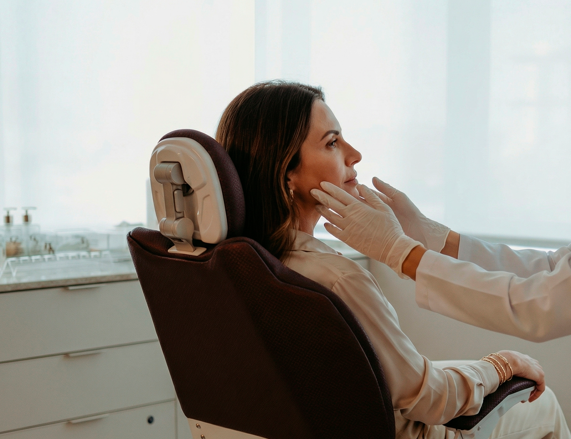 Mulher em uma cadeira de salão durante um tratamento facial, enquanto uma terapeuta aplica produtos em seu rosto.
