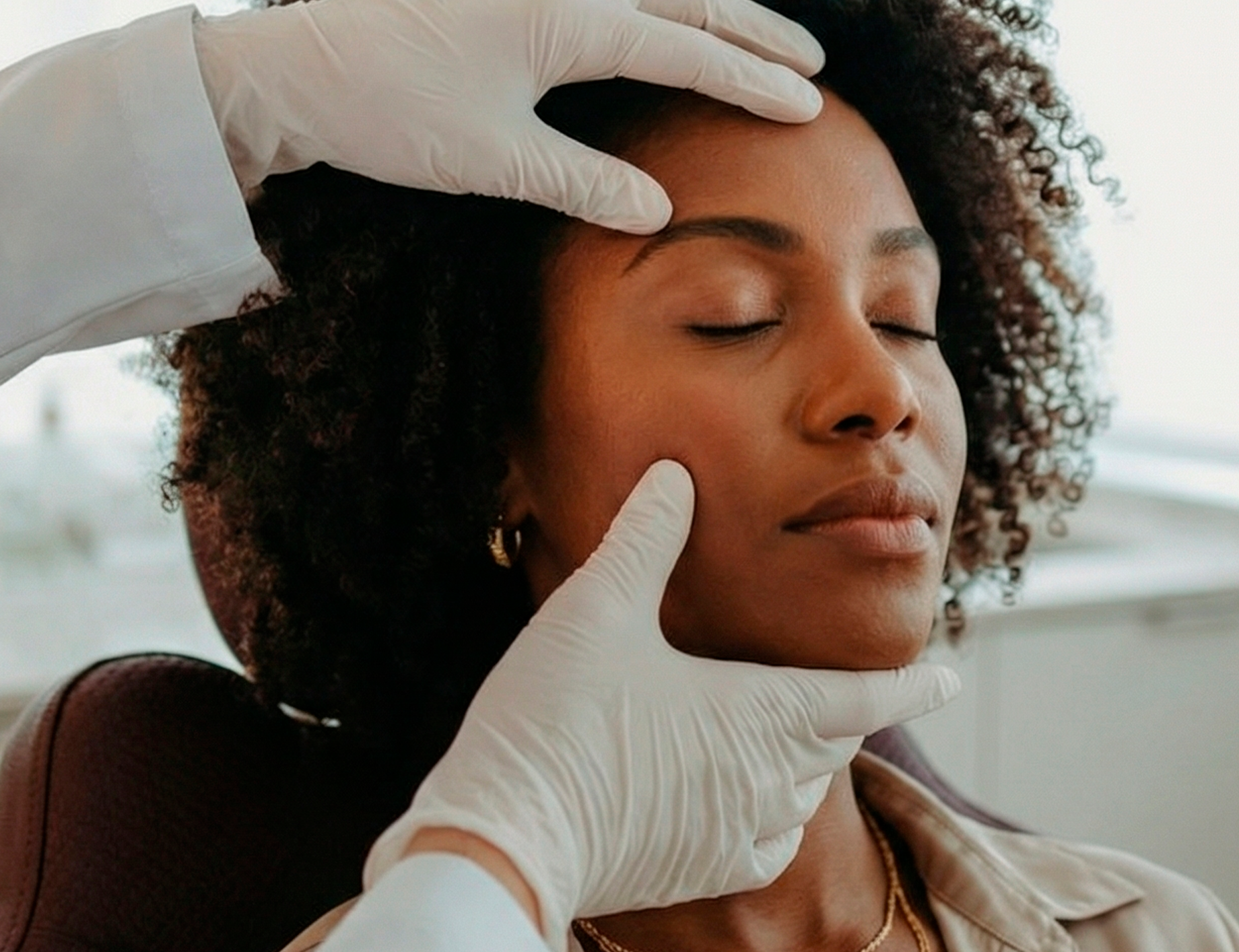 Mãos enluvadas realizando um tratamento facial em uma cliente relaxada com os olhos fechados.
