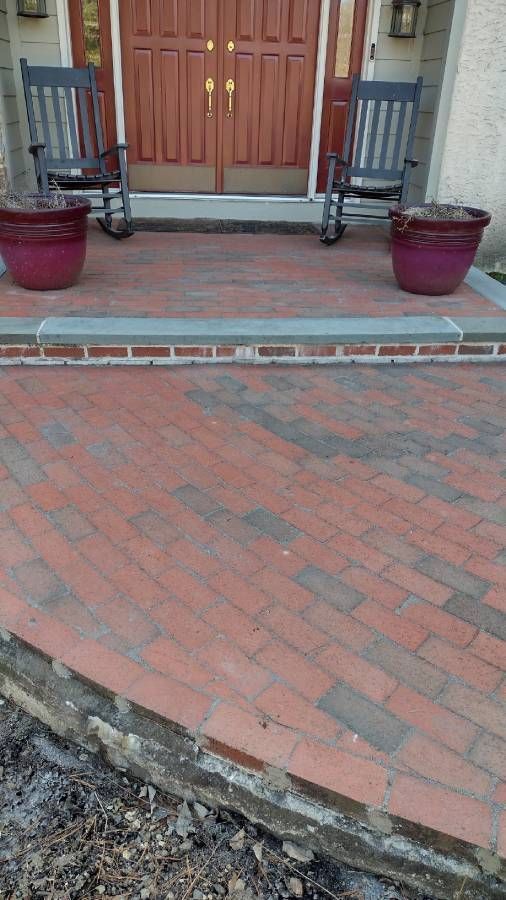 Front porch with red brick walkway, wooden double doors, two black chairs, and potted plants.