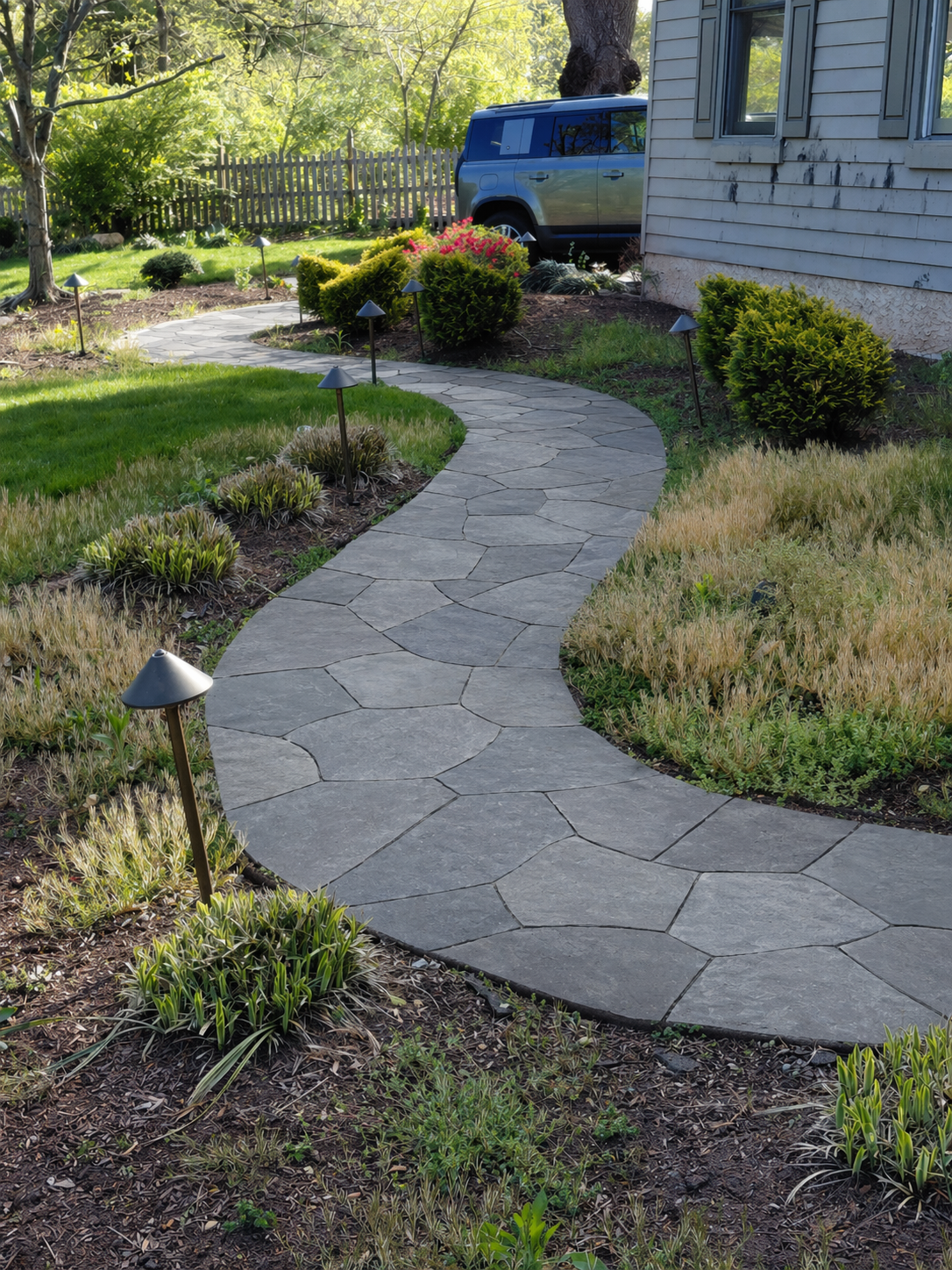 Curved stone garden path through landscaped yard, with shrubs, flowers, and a blue truck near a house.