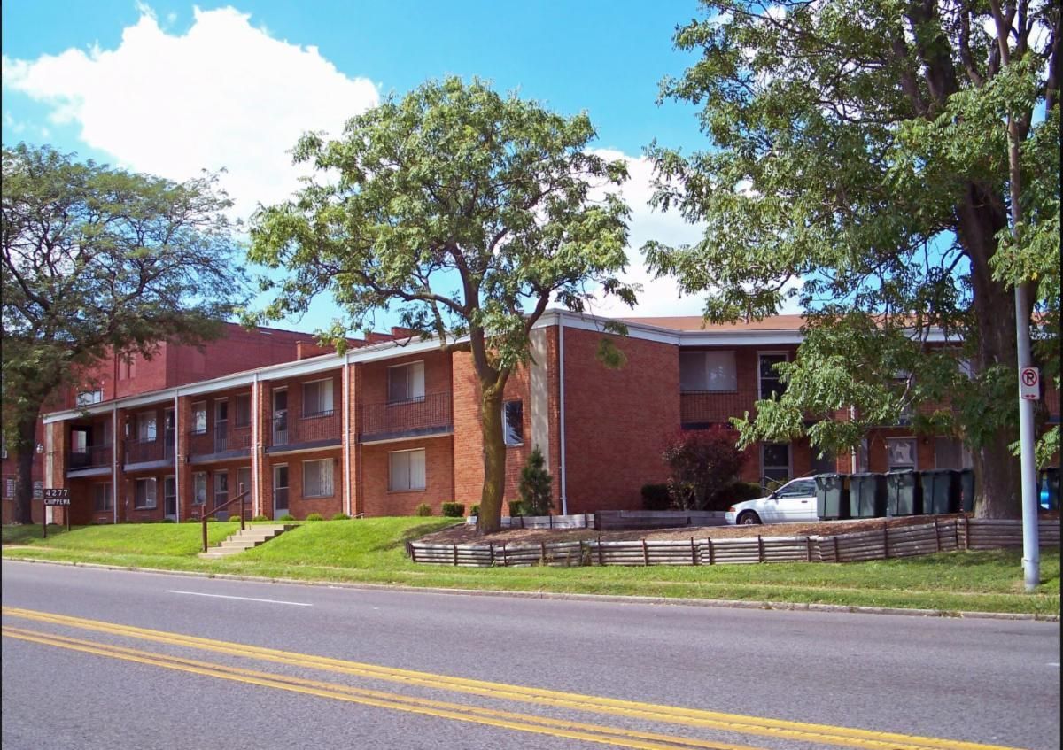 Brick apartment building beside a road, with trees and a bright blue sky.